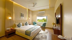 A hotel room with a large bed, a desk with a chair, a TV, and a window with a view of greenery. - THE GANGA RESORT (Bikaner)
