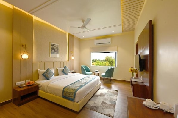 A hotel room with a large bed, a desk with a chair, a TV, and a window with a view of greenery. - THE GANGA RESORT (Bikaner)
