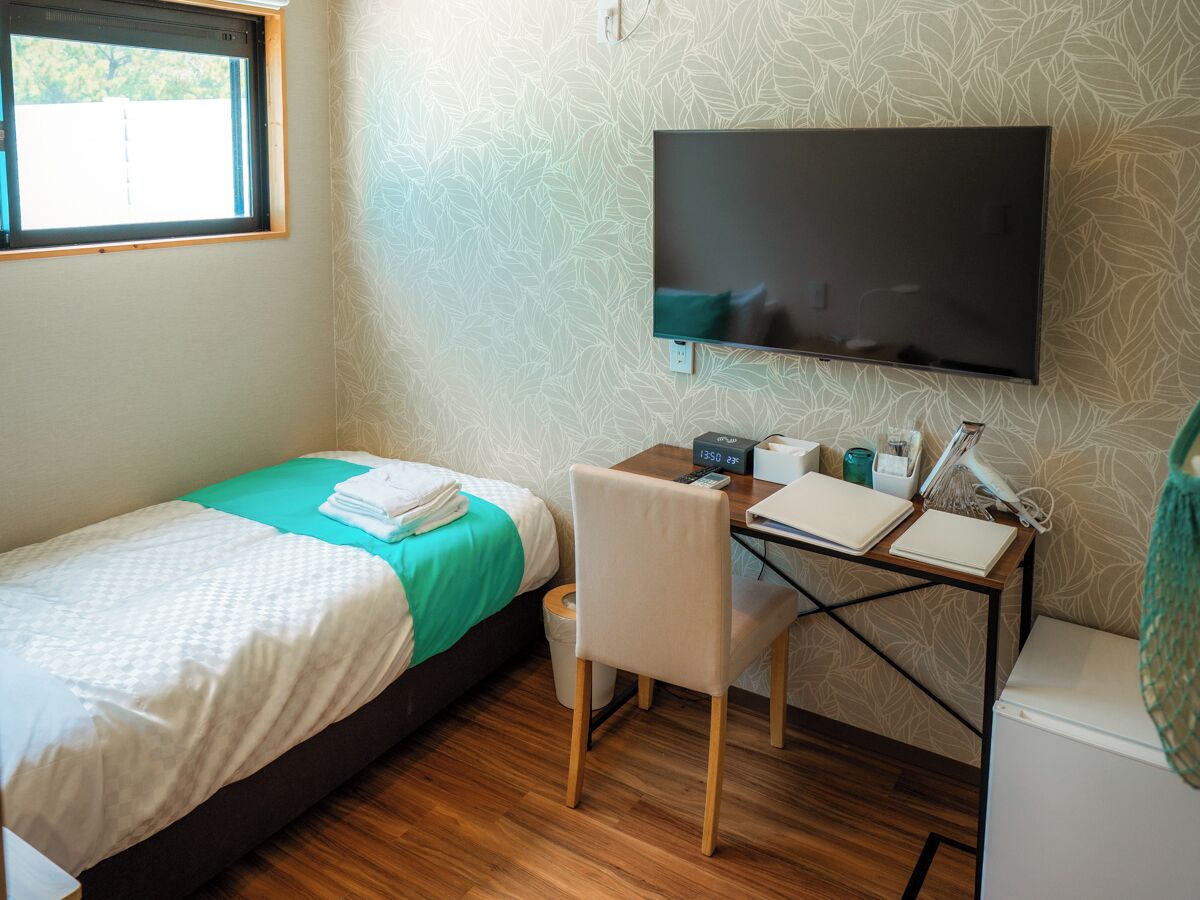 A bedroom with a bed, a desk, a chair, a TV, and a window.