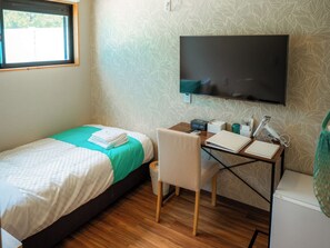A bedroom with a bed, a desk, a chair, a TV, and a window. - MIYAKOJIMA Blue Village HOTEL & SAUNA (Miyakojima)