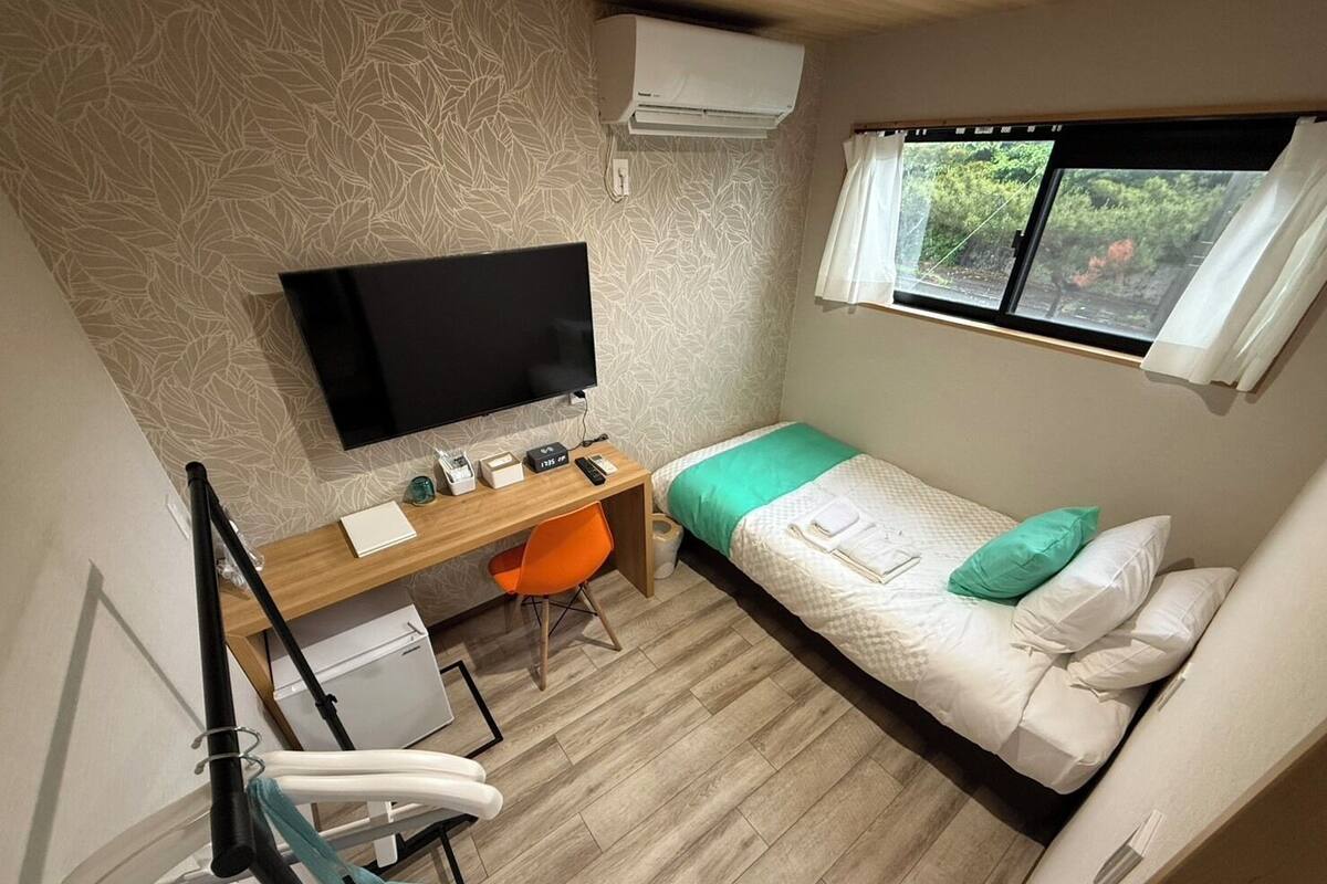 Une pièce de taille modeste comprenant un lit, un bureau avec une chaise, un téléviseur et une fenêtre donnant sur un espace verdoyant.