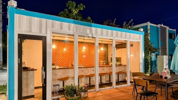 Un espace bar extérieur moderne, doté d’une terrasse en bois, de tabourets de bar et d’une structure en verre avec un mur décoré de motifs.