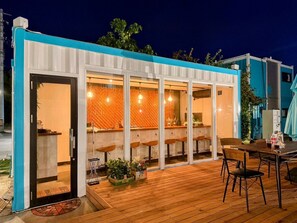 Un espace bar extérieur moderne, doté d’une terrasse en bois, de tabourets de bar et d’une structure en verre avec un mur décoré de motifs.