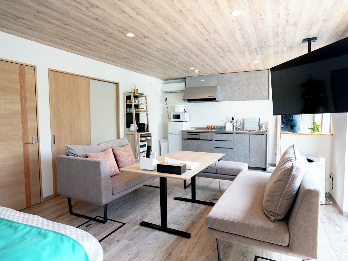 A modern living room with a sofa, a coffee table, and a TV. There is a kitchen area with cabinets and a microwave.