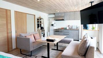 A modern living room with a sofa, a coffee table, and a TV. There is a kitchen area with cabinets and a microwave.