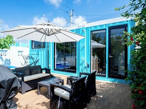 Un patio al aire libre con mobiliario de mimbre, un paraguas grande y vistas a una piscina a través de puertas de cristal.