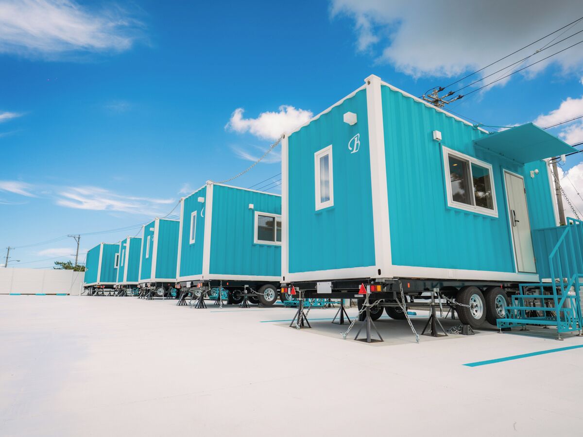A row of blue mobile homes with white trim, each with a single window and a door, parked on a flat surface with a clear sky in the background.