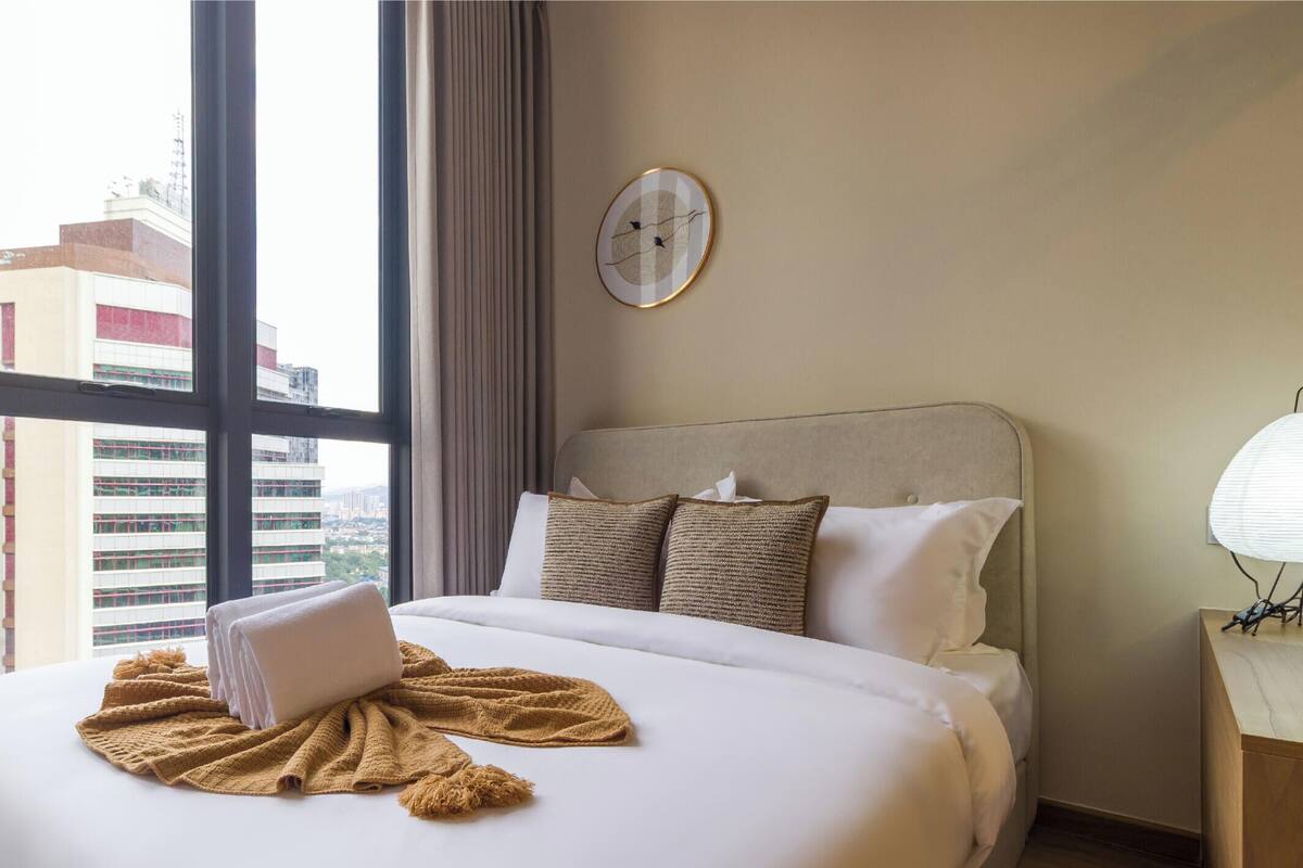 A hotel room with a large bed, a view of buildings through the window, and a wall clock.