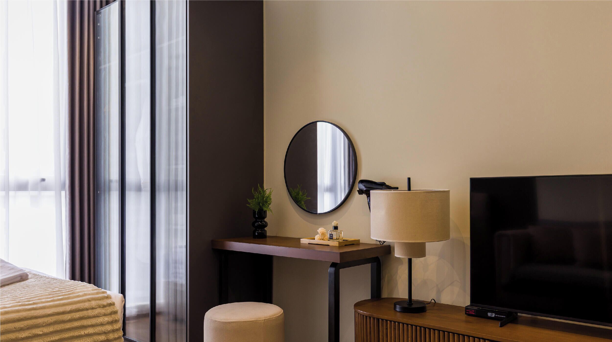 A hotel room with a bed, a TV, a desk with a lamp, a mirror, and a window with curtains.