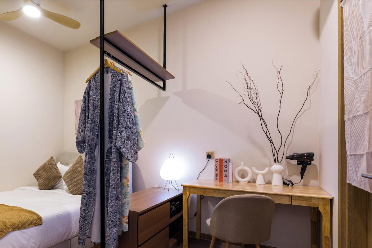 A bedroom with a bed, a desk, a chair, a hanging robe, a lamp, and a wall-mounted hairdryer.