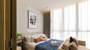 Ein modernes Hotelzimmer mit einem Bett, einem Nachttisch mit Pflanze, einem gerahmten Kunstwerk an der Wand und einem Fenster mit Vorhängen.