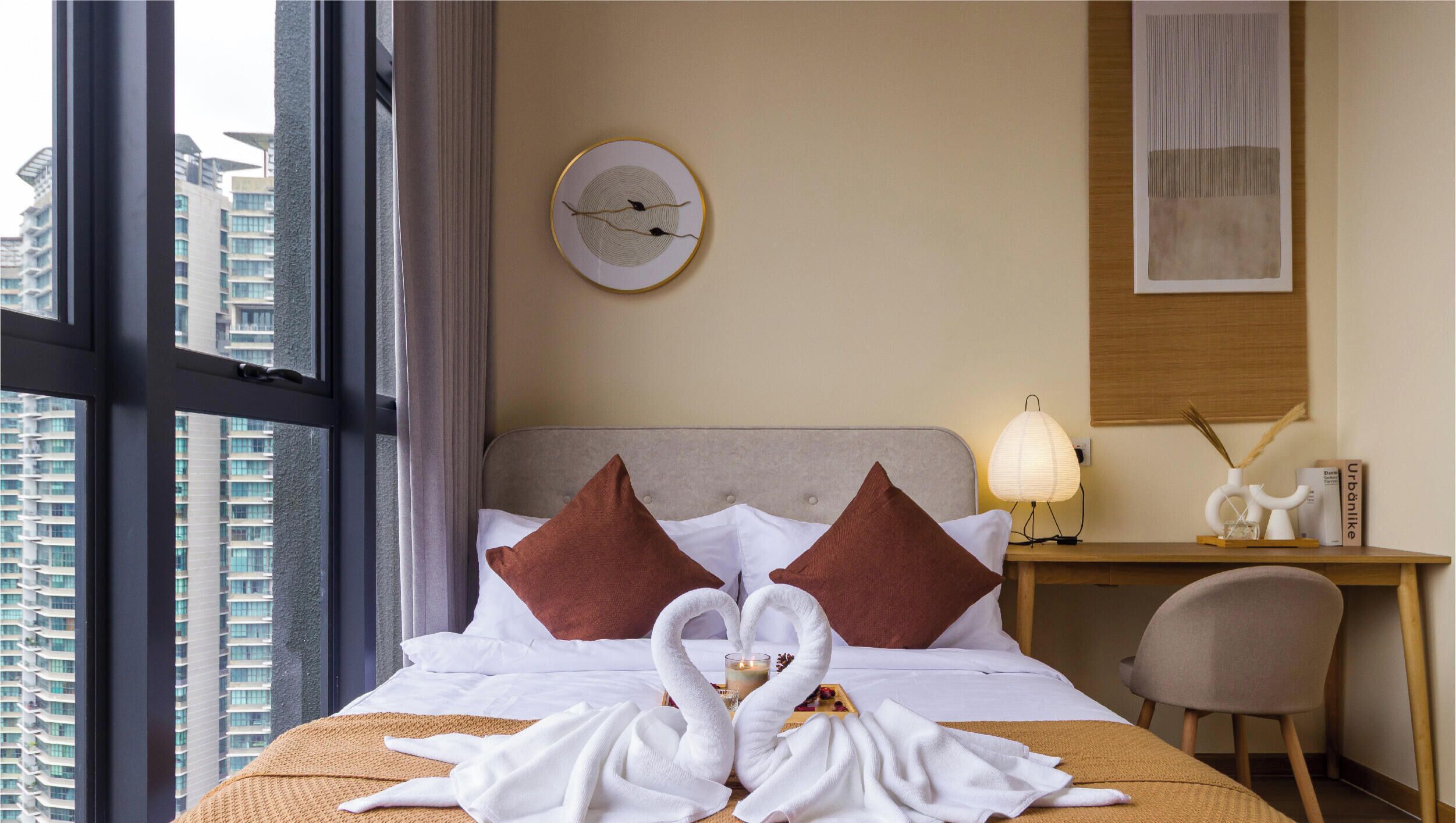 A hotel room with a bed, a desk, a chair, a clock on the wall, and a view of buildings through the window.