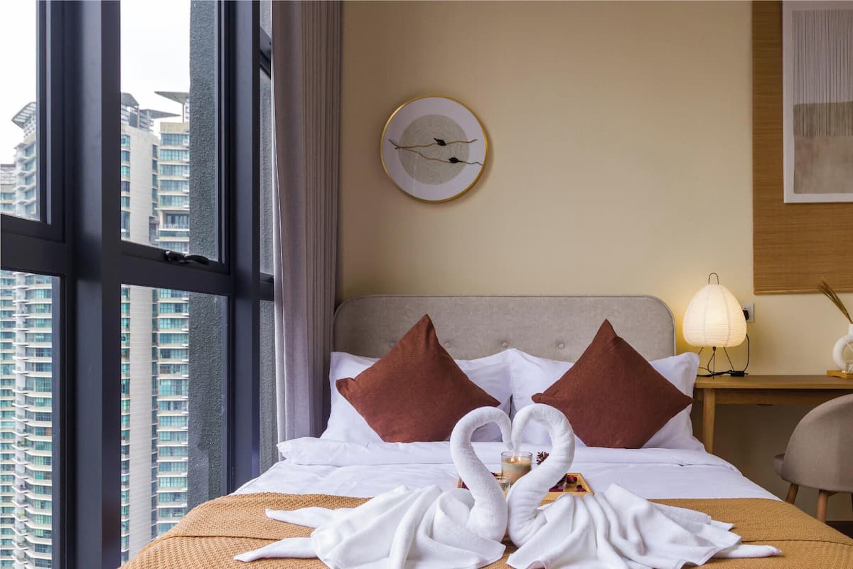 A hotel room with a bed, a desk, a chair, a clock on the wall, and a view of buildings through the window.