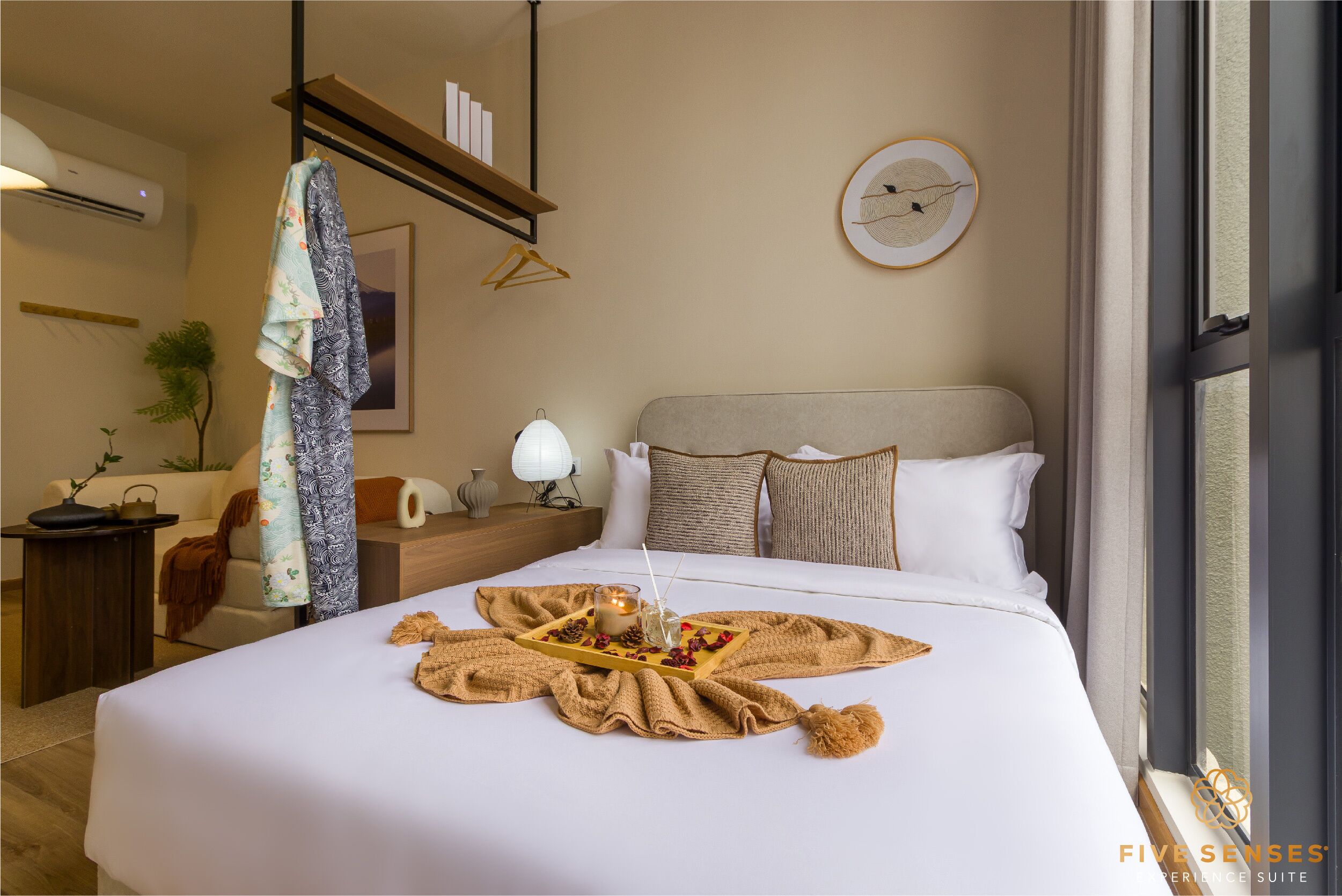 A hotel room with a neatly made bed, a bedside table with a lamp, a hanging coat, and a framed picture on the wall.