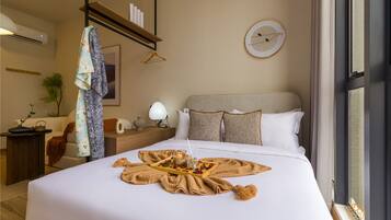 A hotel room with a neatly made bed, a bedside table with a lamp, a hanging coat, and a framed picture on the wall.
