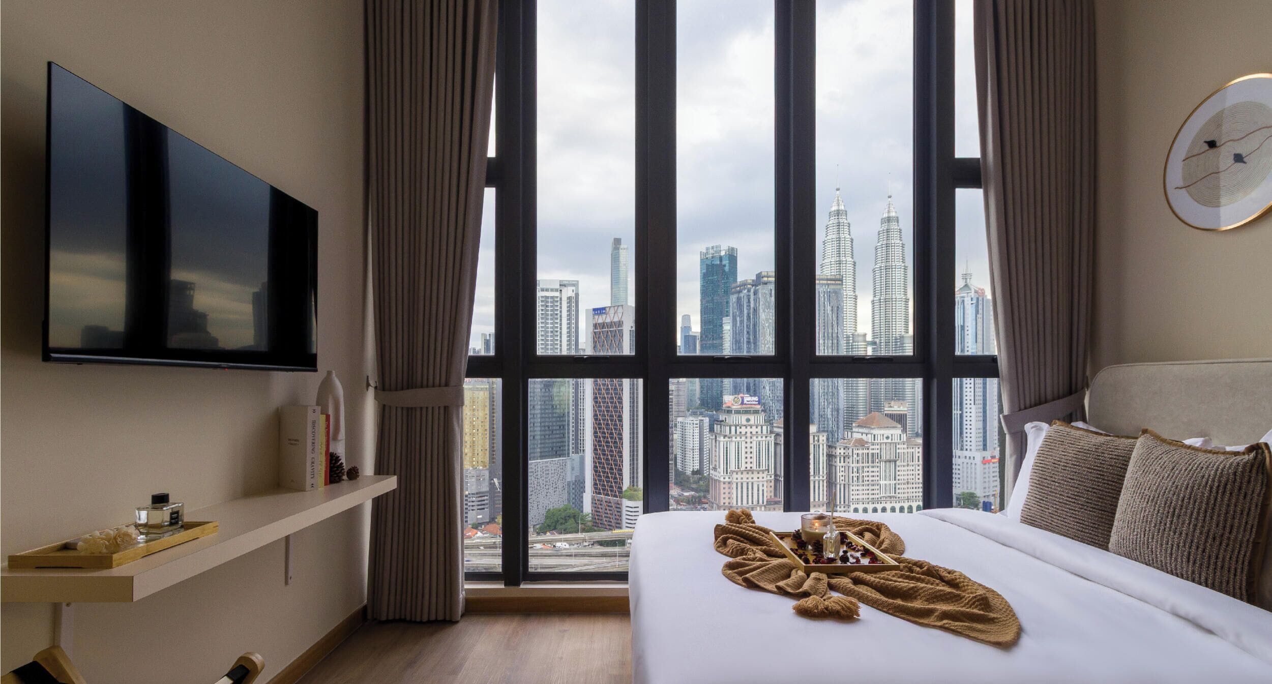 Une chambre d’hôtel dotée d’une grande fenêtre donnant sur la ville, d’un téléviseur à écran plat fixé au mur, d’une table de chevet avec un plateau, et d’un lit avec une couverture et des oreillers.