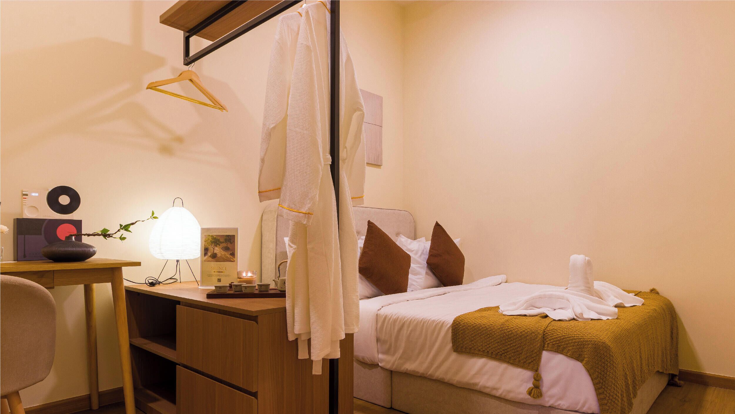 A hotel room with a bed, a desk, a chair, a hanging towel, and a bedside table with a lamp and a record player.