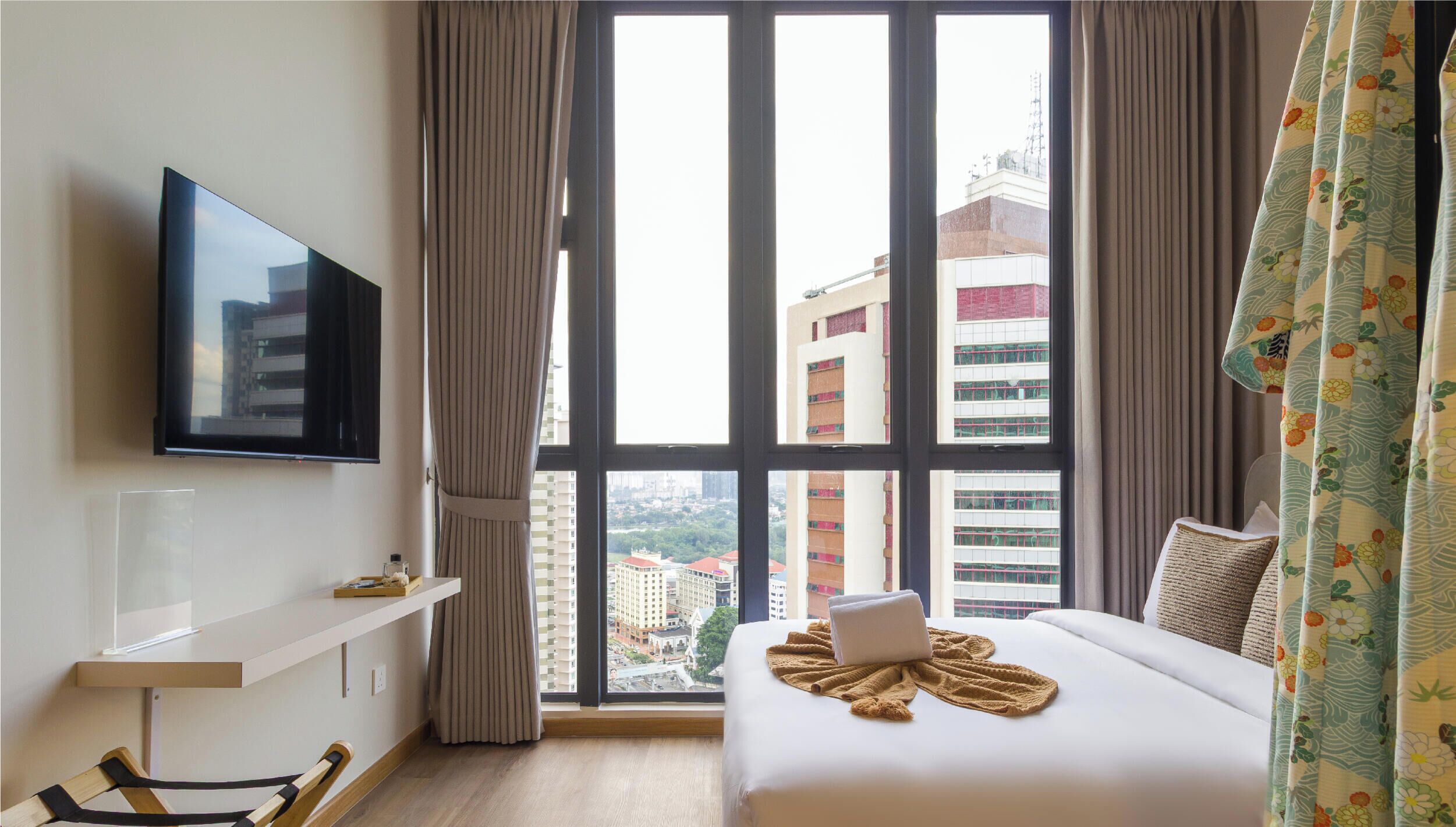 Habitación de hotel con un ventanal grande, televisor de pantalla plana, una cama con una laptop y un estante con un objeto pequeño.