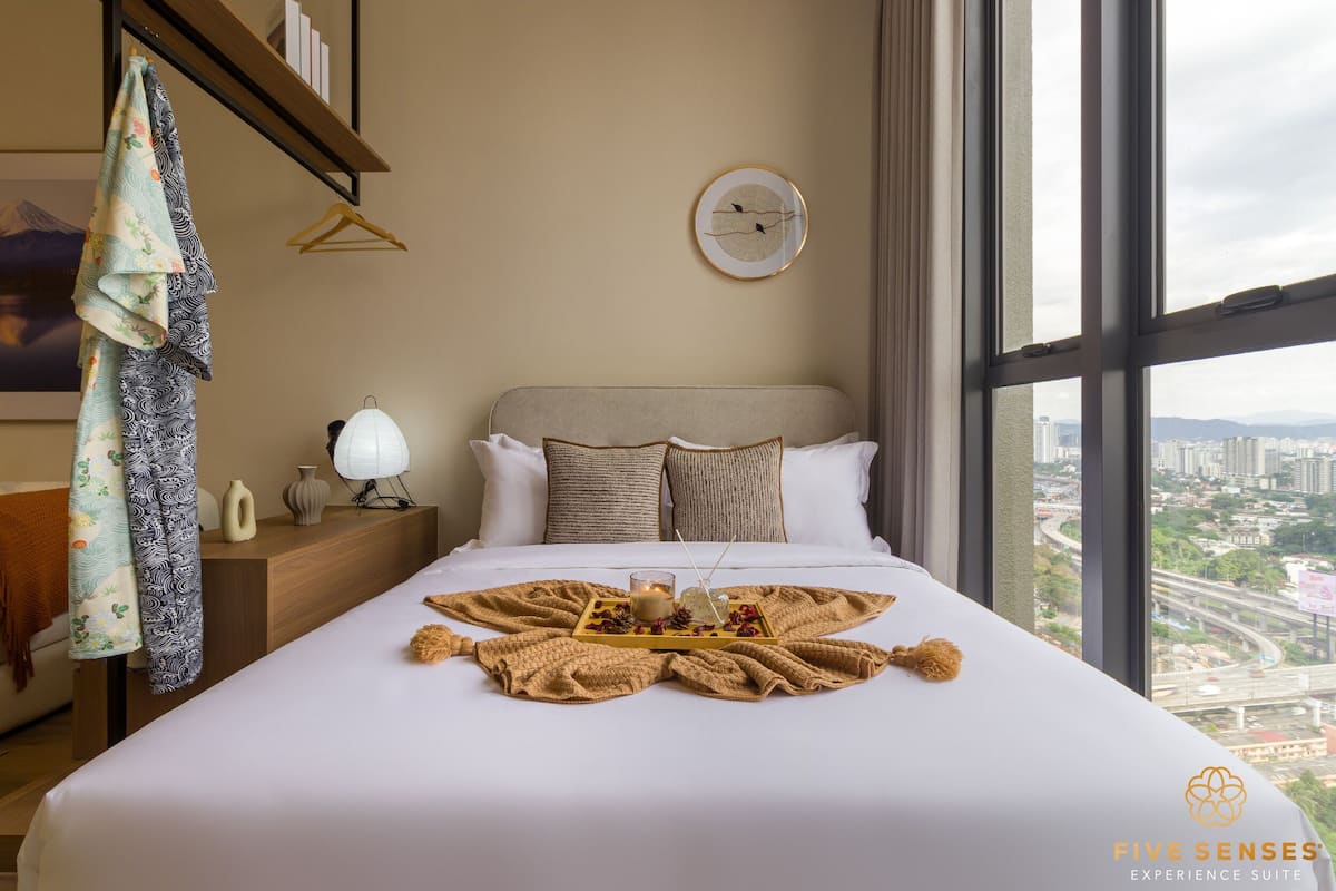 A neatly made bed with a tray of snacks and a candle, a clock on the wall, and a city view through large windows.