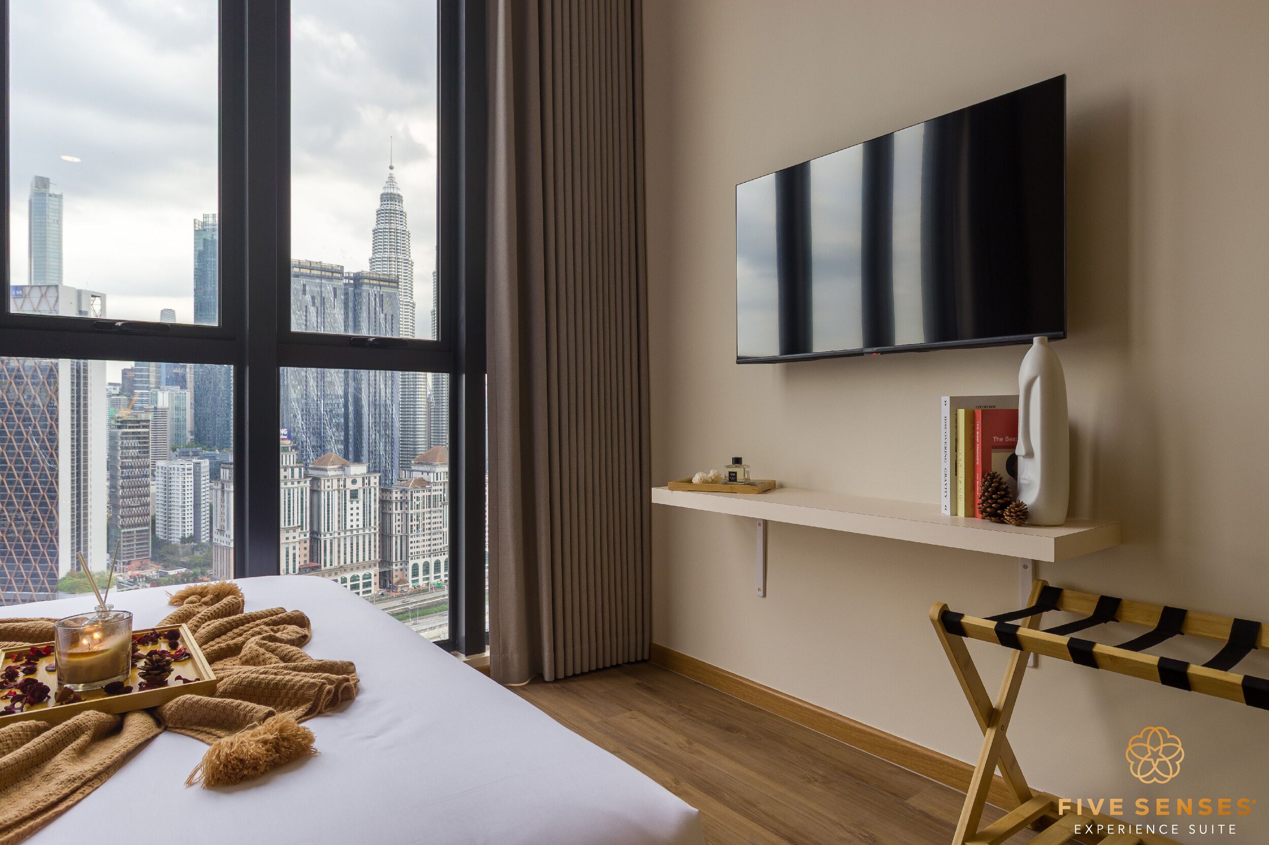 A modern hotel room with a large window, a bed with a view of the city skyline, a mounted TV, a bedside table with books and a vase, and a wooden side table.