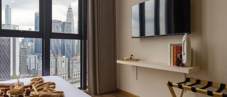 A modern hotel room with a large window, a bed with a view of the city skyline, a mounted TV, a bedside table with books and a vase, and a wooden side table.