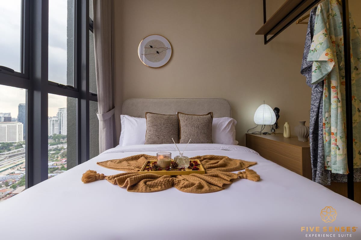 A hotel room with a bed, a bedside table with a lamp, a clock on the wall, and a view of the city through large windows.