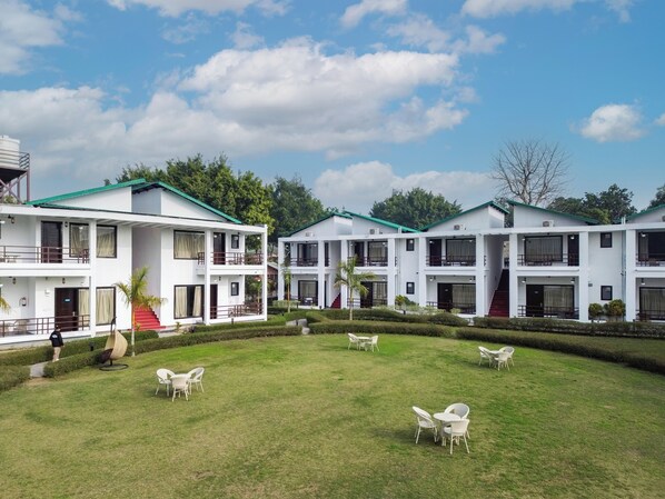 A white building with multiple floors and balconies, surrounded by a well-maintained lawn with white chairs and tables. - Baris resort Corbett (Ramnagar)