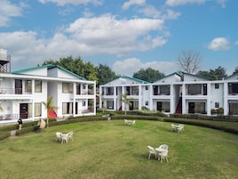 Un bâtiment blanc à étages, doté de nombreux balcons, entouré d’une pelouse bien entretenue où sont disposées des chaises et des tables blanches.