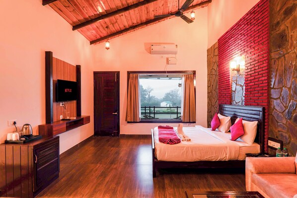 A hotel room with a wooden ceiling, a bed with pink and white bedding, a TV on the wall, and a stone wall. - Baris resort Corbett (Ramnagar)