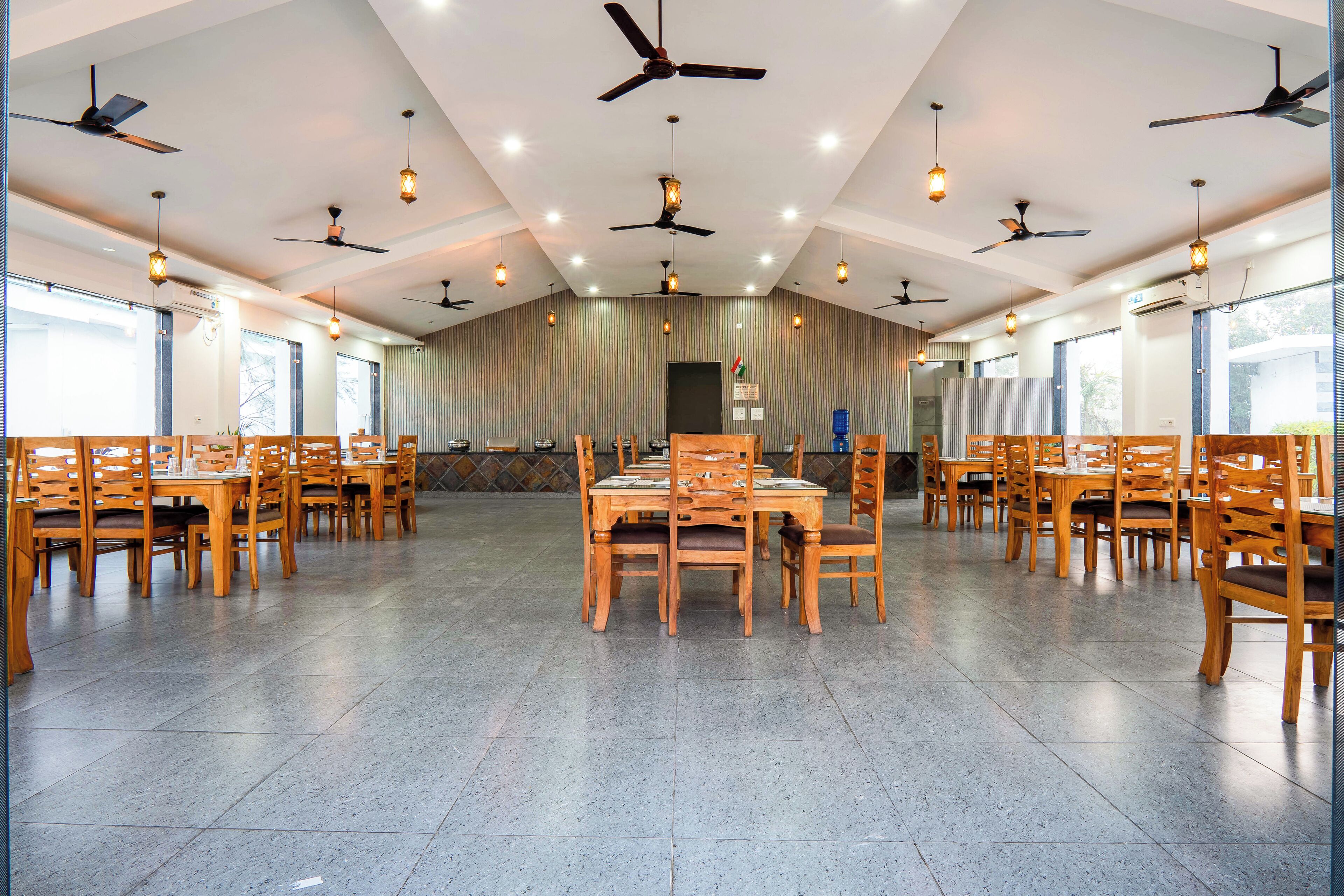 A spacious dining hall with multiple wooden tables and chairs, ceiling fans, and large windows.