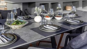 En utomhusplats för måltider med en stenmur, en grill och ett bord dukat för sex personer med tallrikar, glas och bestick.