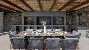 An outdoor dining area with a stone wall backdrop, a large table set for six, and wicker chairs. - Celestial II             ., Villa (Paros)