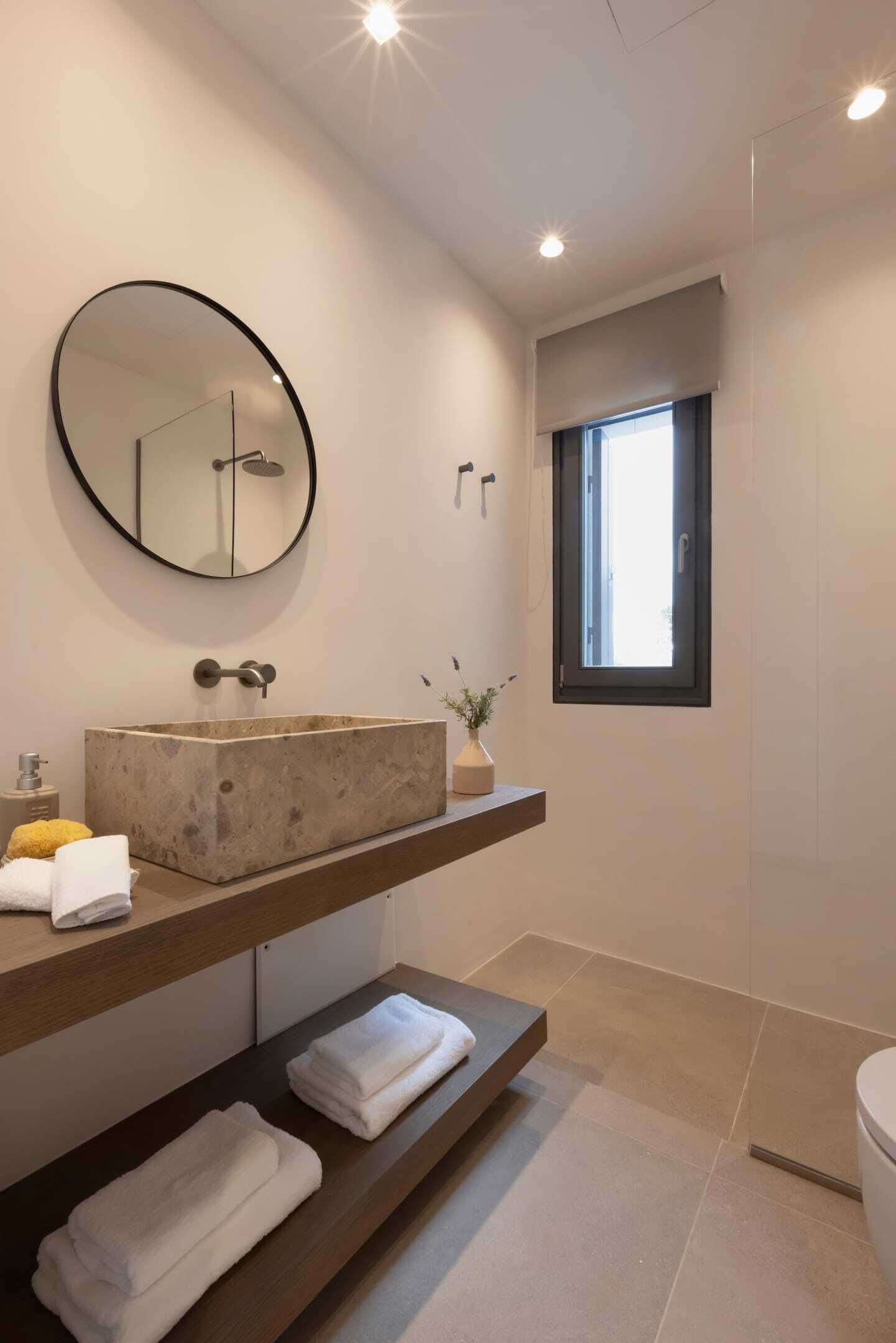 Un baño moderno con un espejo grande y redondo, un lavamanos rectangular y una ventana.