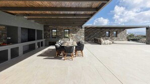 An outdoor patio with a stone fireplace, a dining table with chairs, and a seating area with a sofa and a coffee table. - Celestial II             ., Villa (Paros)