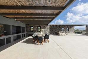 Eine Terrasse im Freien mit einem Kamin aus Stein, einem Esstisch mit Stühlen und einer Sitzecke mit einer Couch und einem Couchtisch.