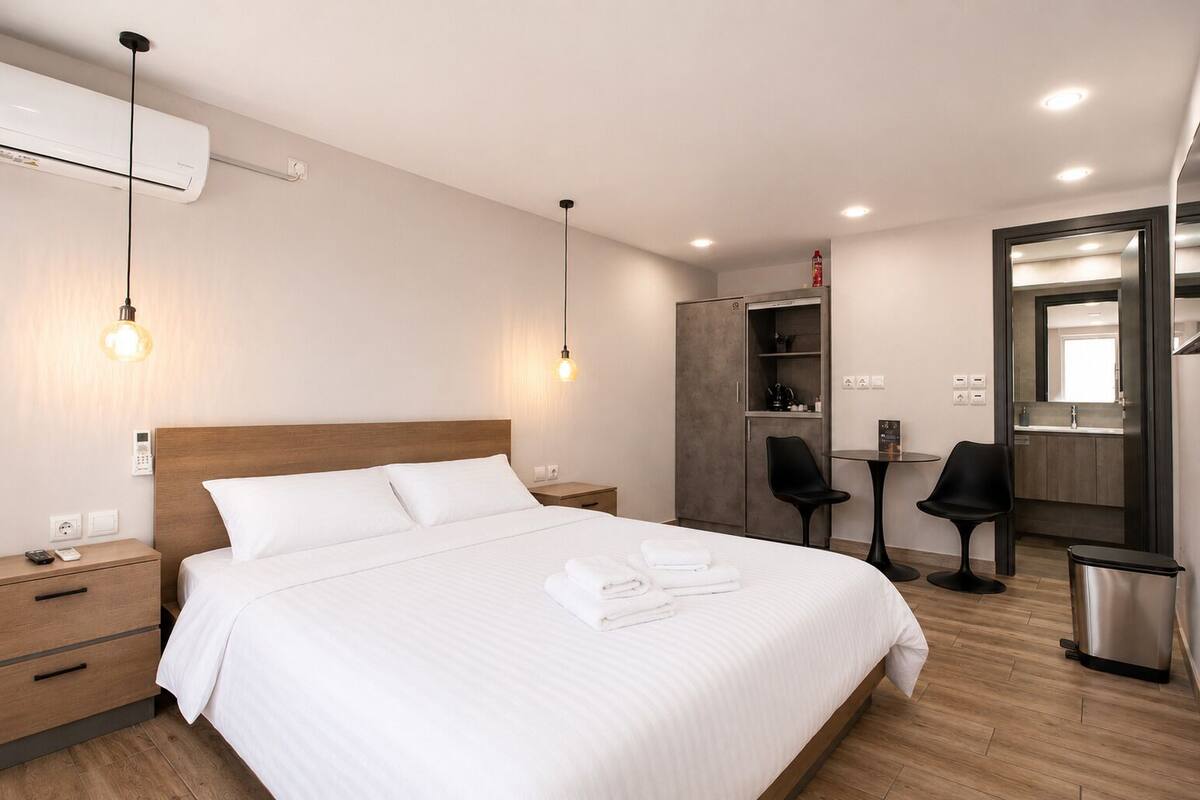 A hotel room with a bed, bedside tables, a wardrobe, a desk with chairs, and a trash bin.