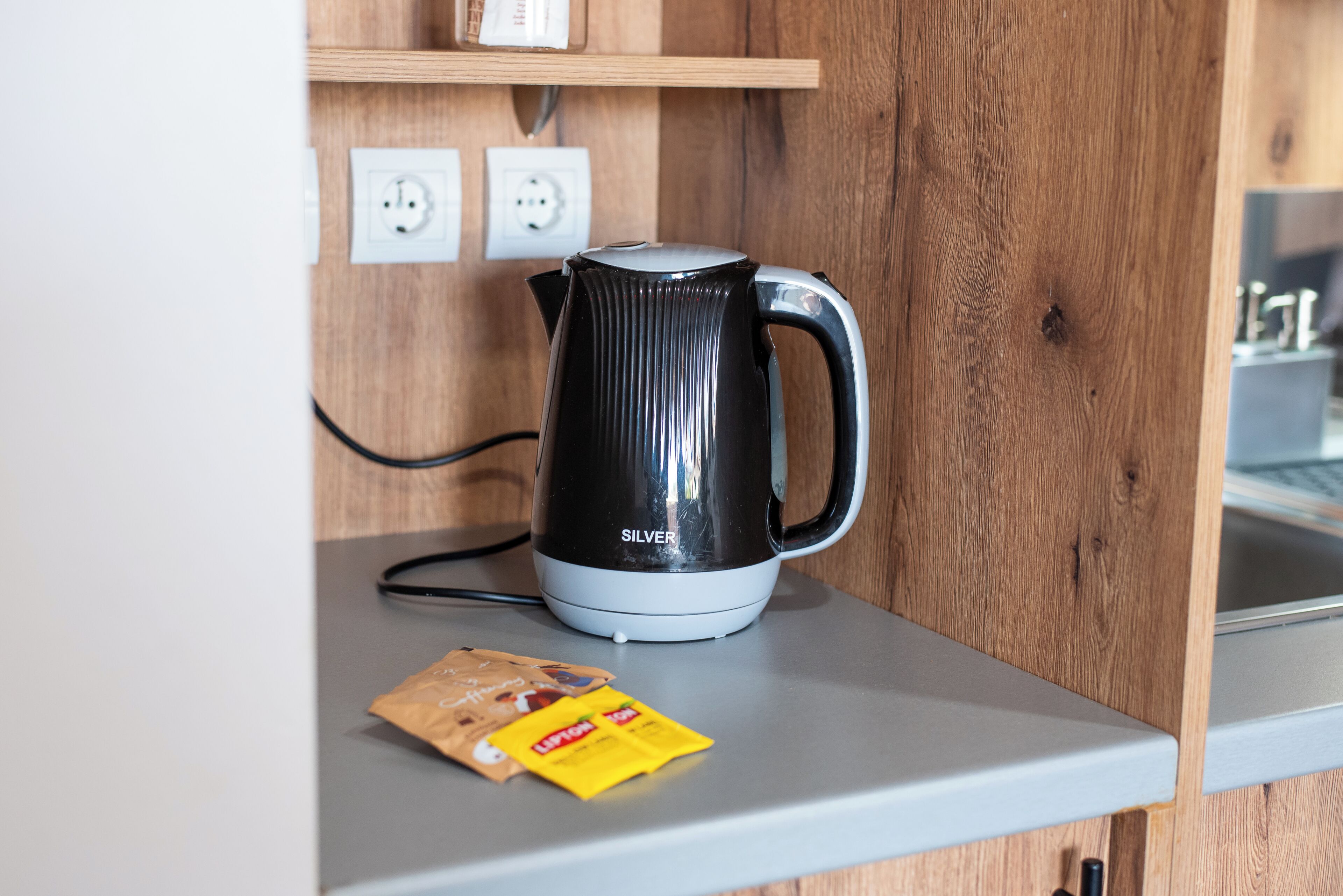 Una zona de cocina con un hervidor blanco y negro sobre una encimera, una cápsula de café y un estante de madera arriba con enchufes eléctricos.