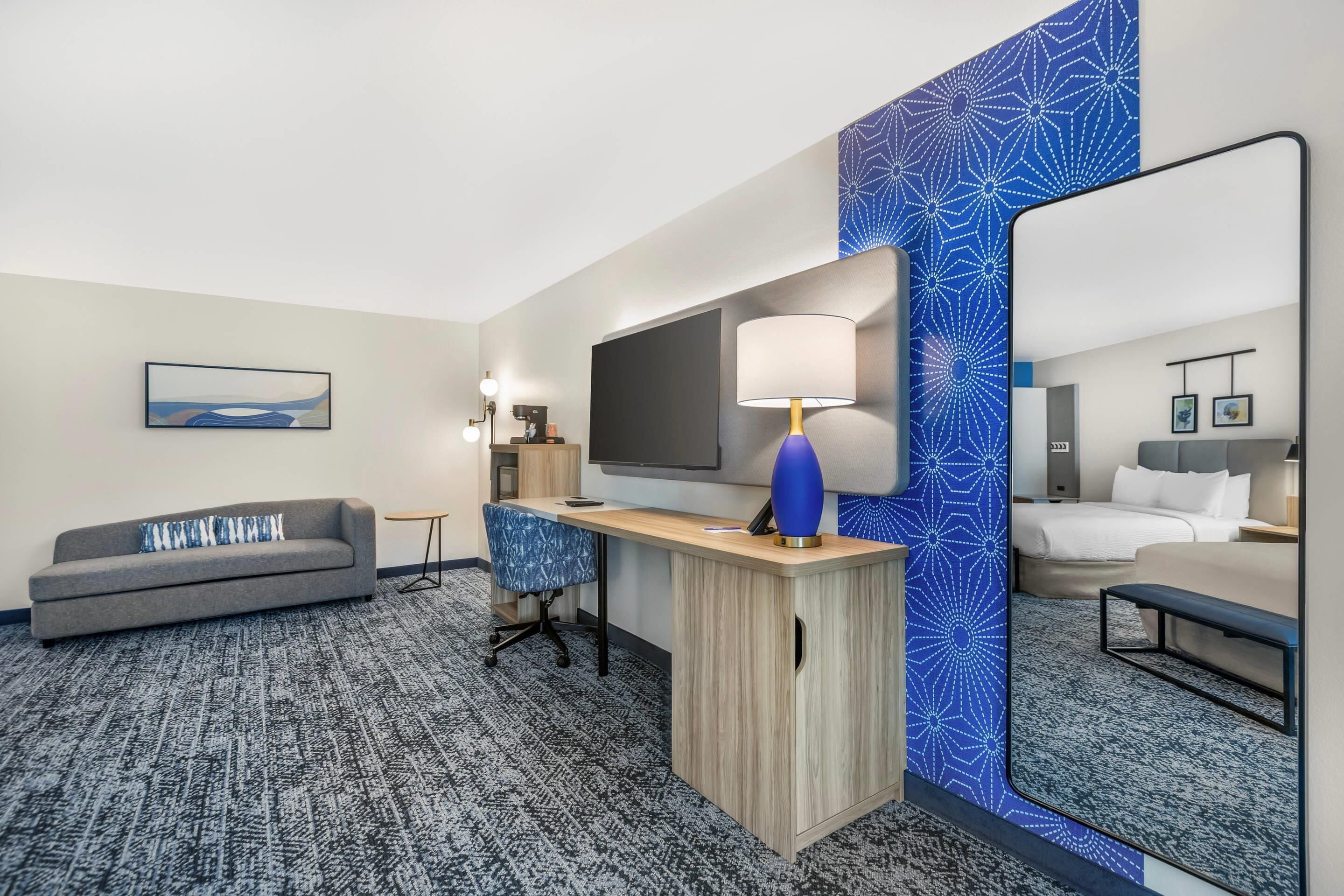A hotel room with a sofa, desk, TV, and a large mirror.