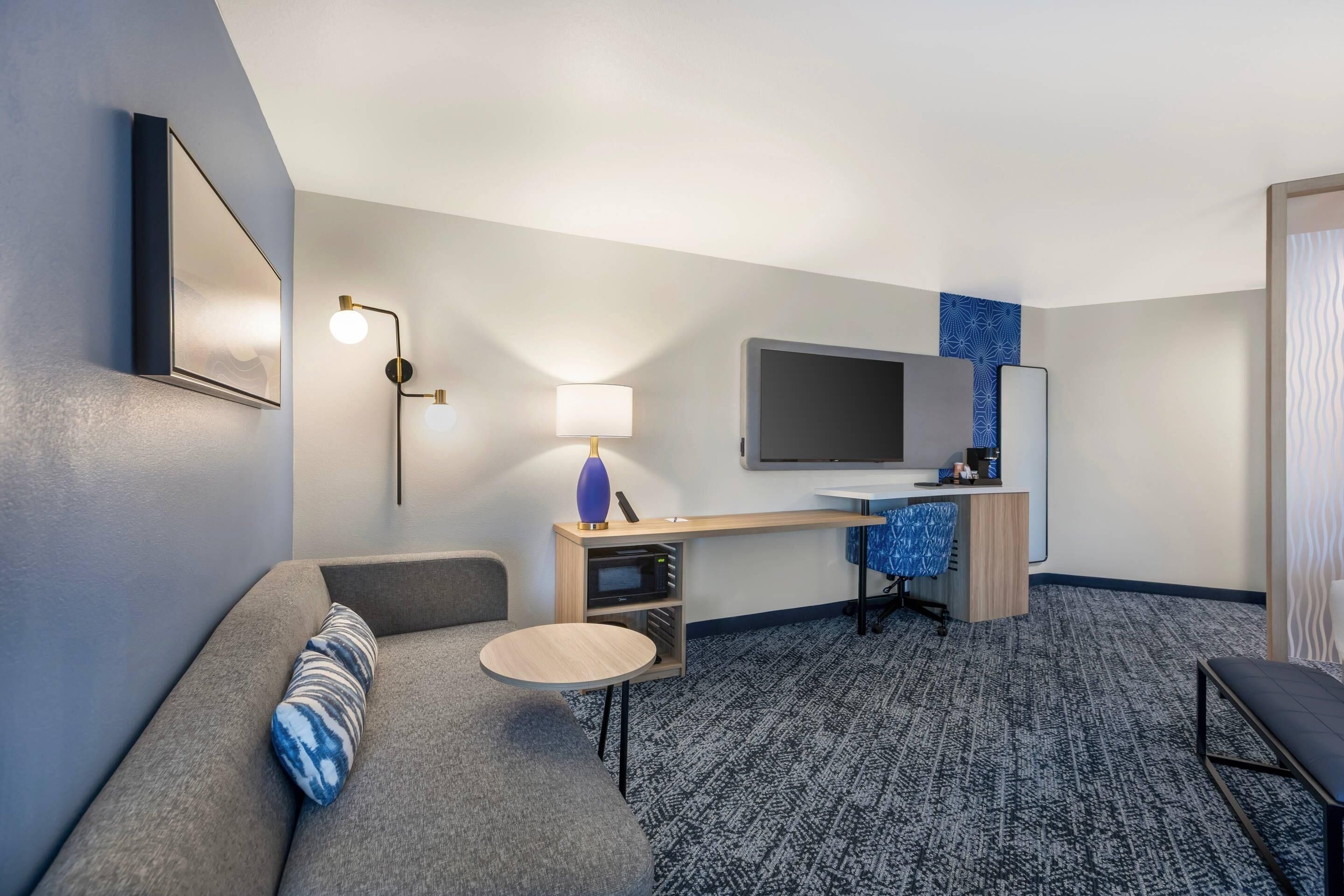 A hotel room with a sofa, a small round table, a desk with a computer and a flat-screen TV, a lamp, and a patterned carpet.