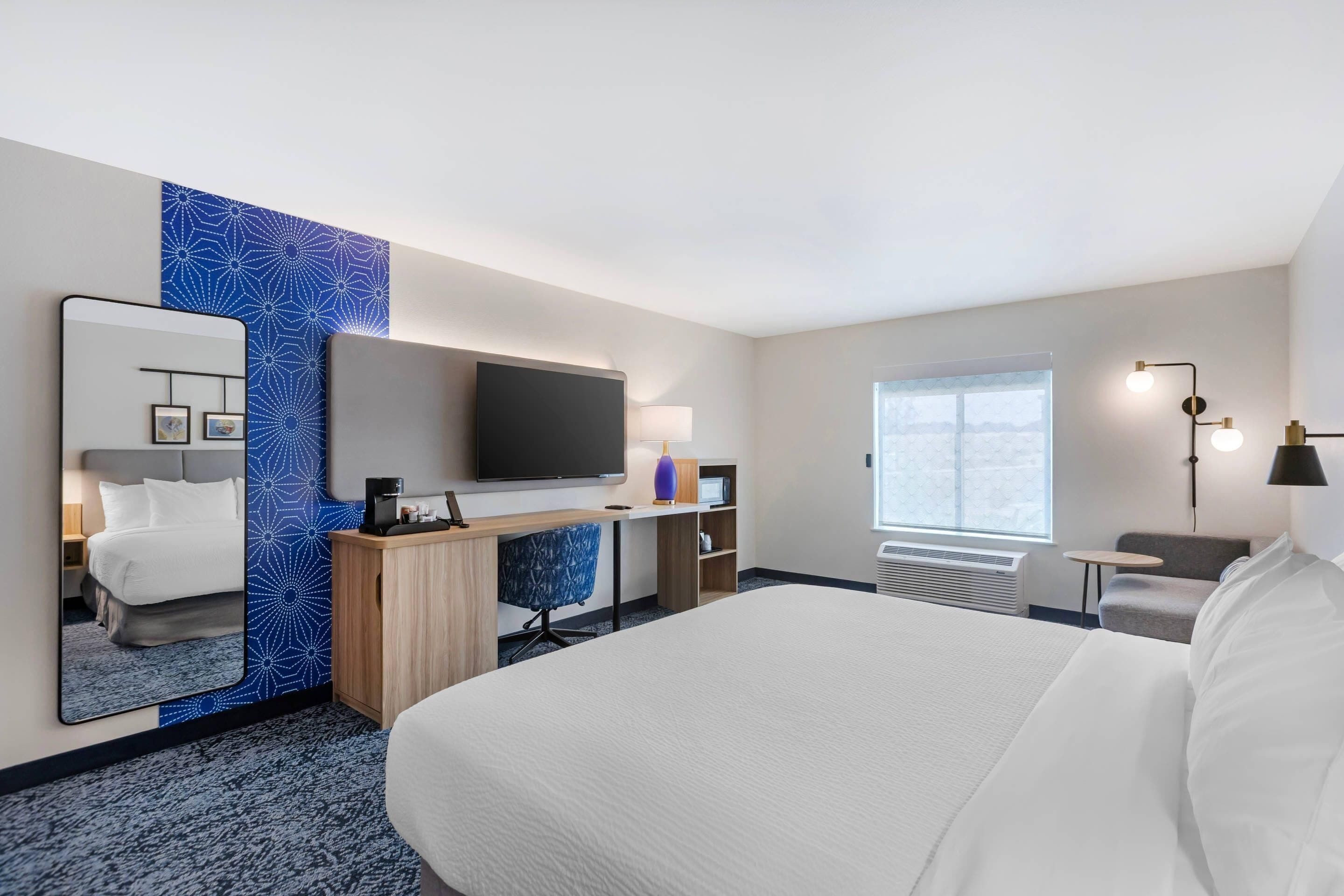 Ein Hotelzimmer mit einem großen Bett, einem Schreibtisch mit Computer, einem Fernseher, einem Spiegel, einem Fenster und einem kleinen Tisch.