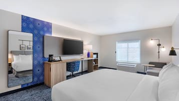 Ein Hotelzimmer mit einem großen Bett, einem Schreibtisch mit Computer, einem Fernseher, einem Spiegel, einem Fenster und einem kleinen Tisch.