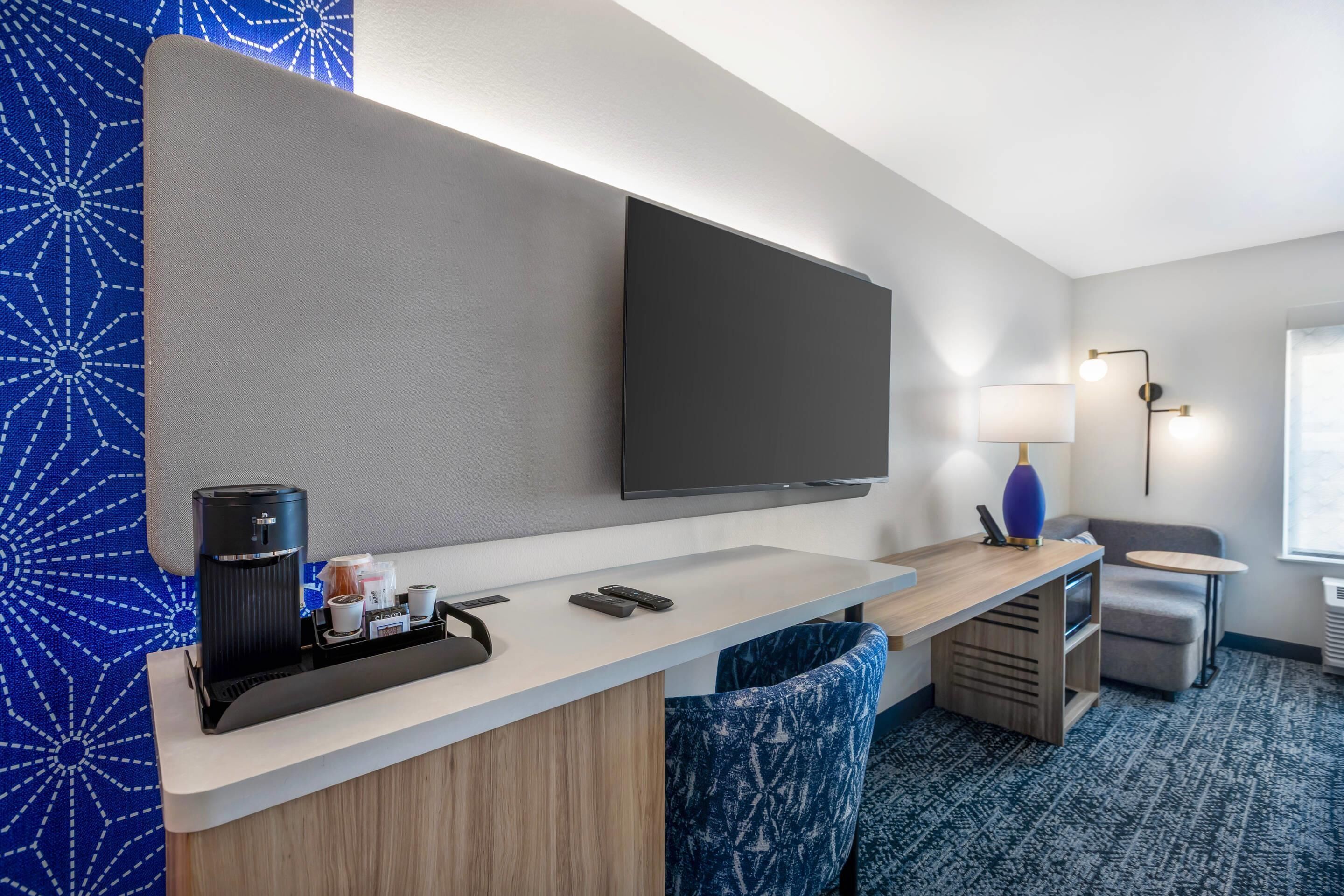 A hotel room with a flat-screen TV mounted on the wall, a coffee machine, a small table with a lamp, a sofa, and a patterned carpet.