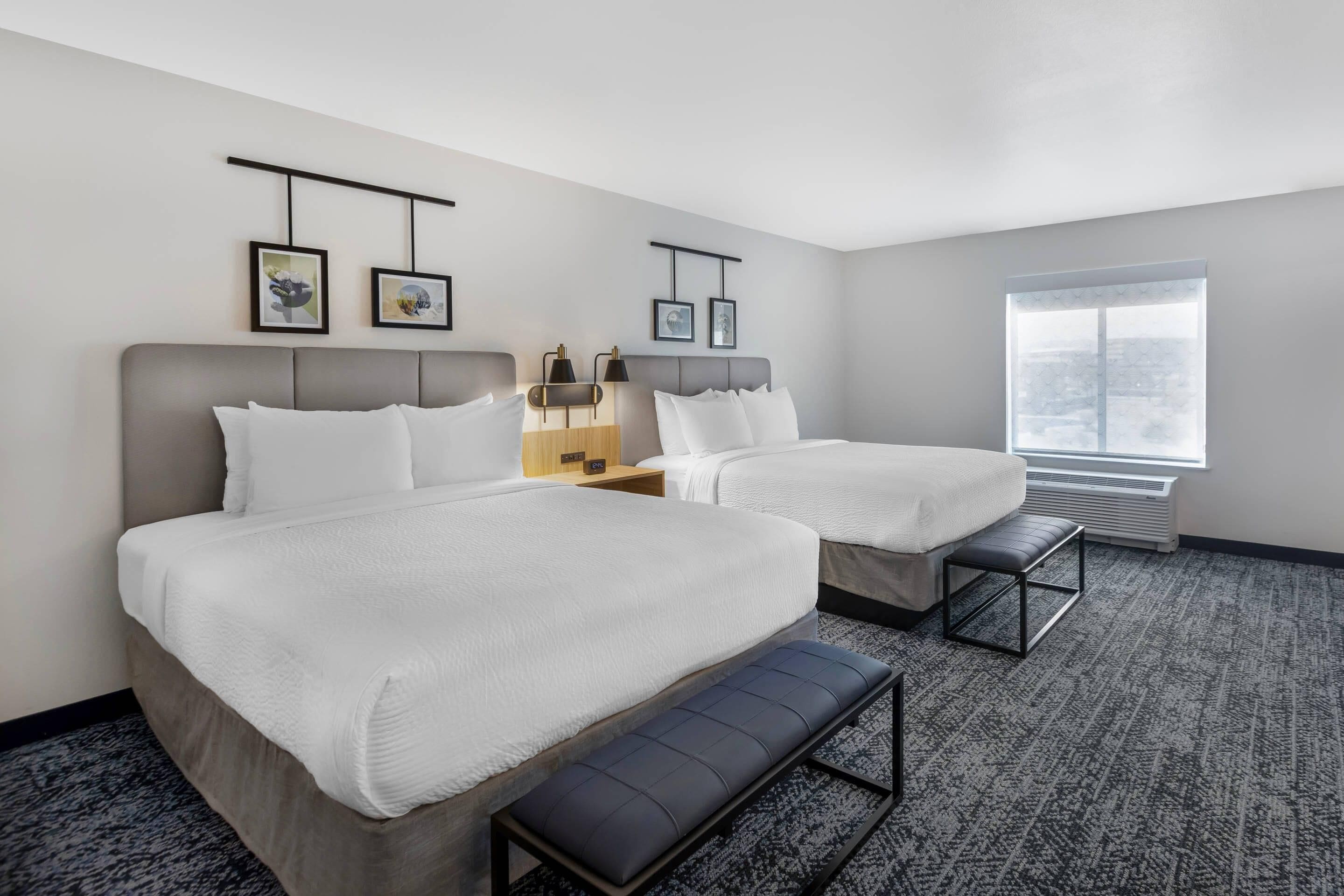 A hotel room with two beds, a bench, a nightstand, a window, and framed pictures on the wall.