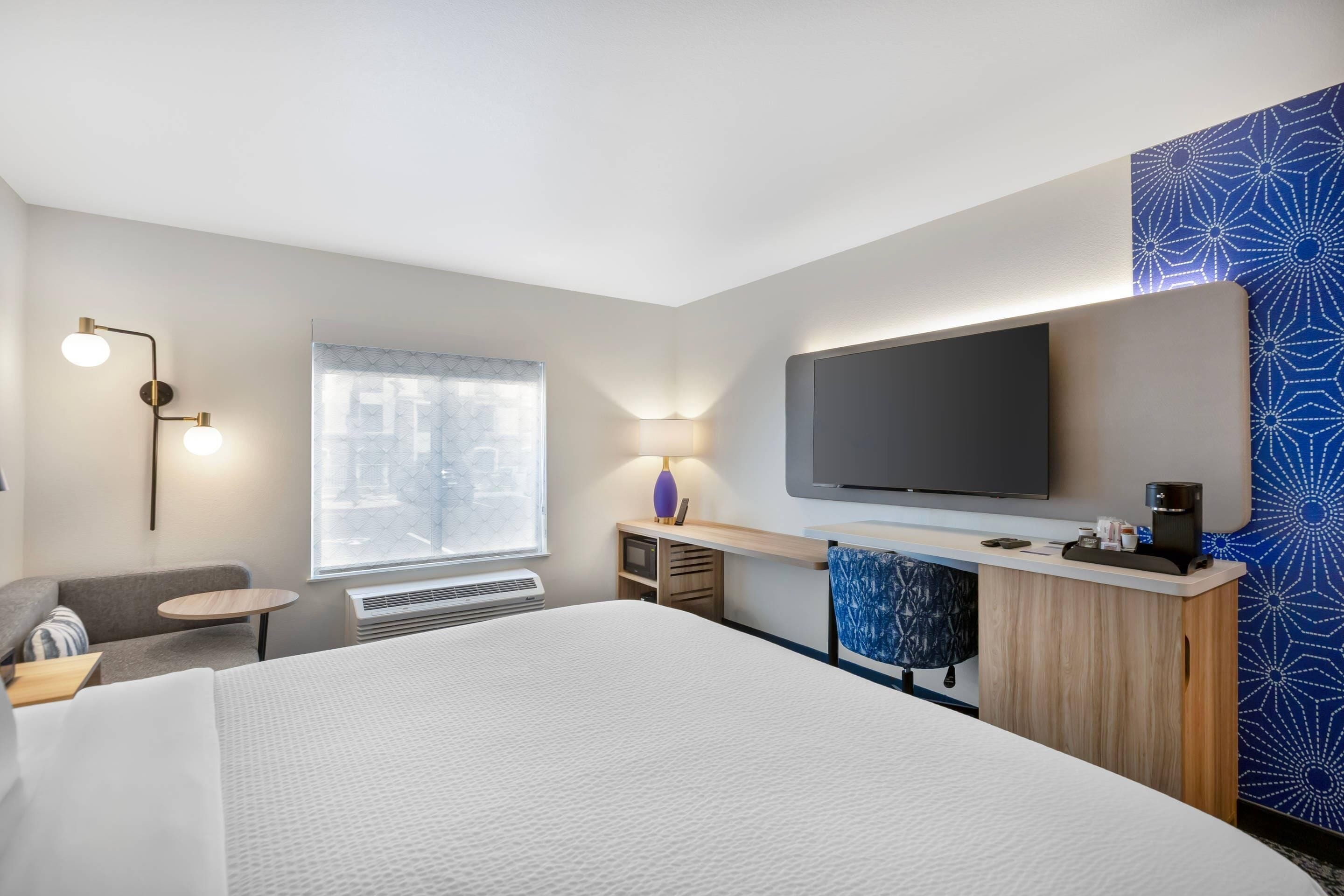A hotel room with a bed, a desk with a flat-screen TV, a chair, a small table, and a window with blinds.