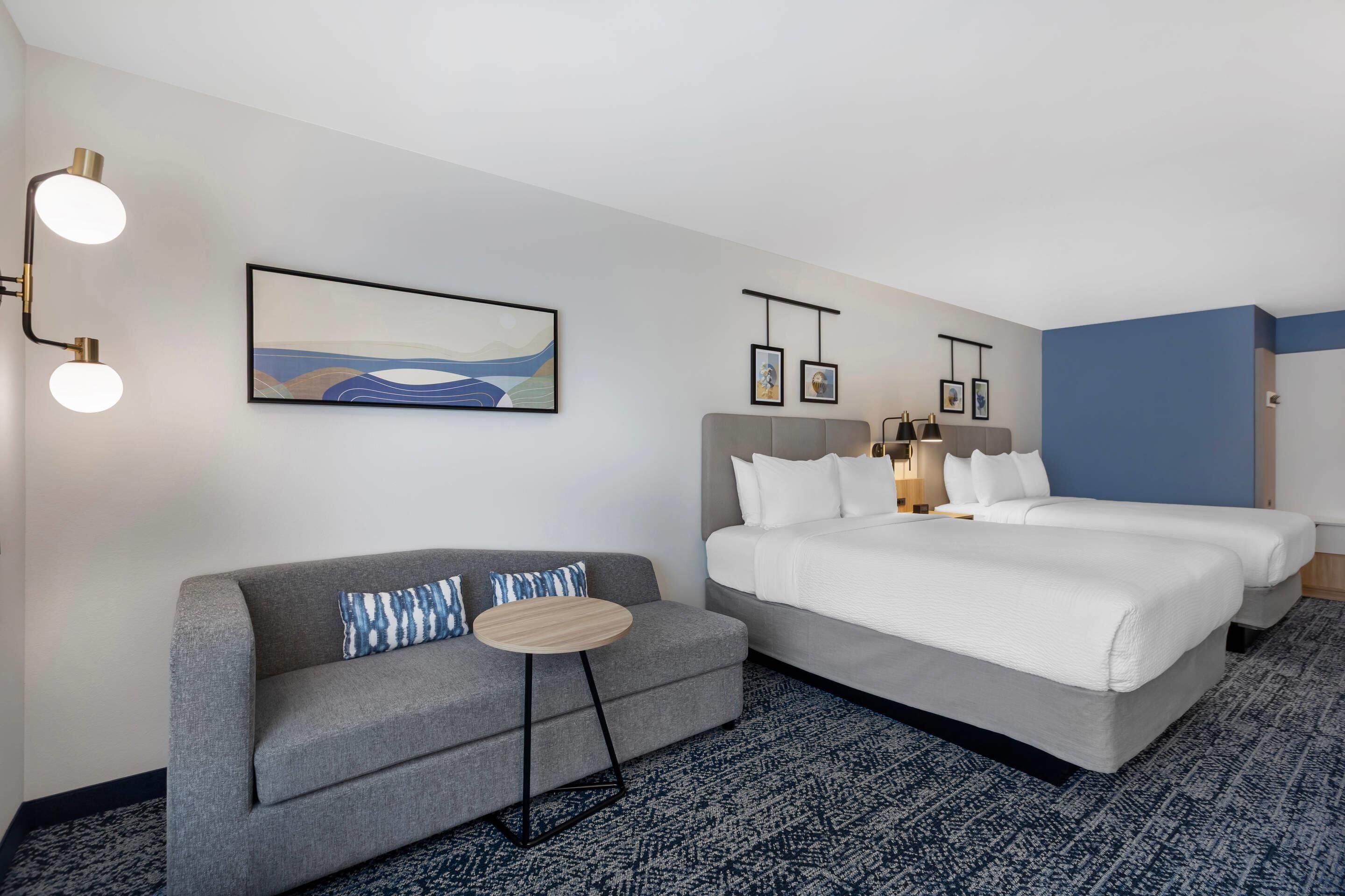 A hotel room with a grey sofa, a round coffee table, a bed with white bedding, and a blue accent wall.