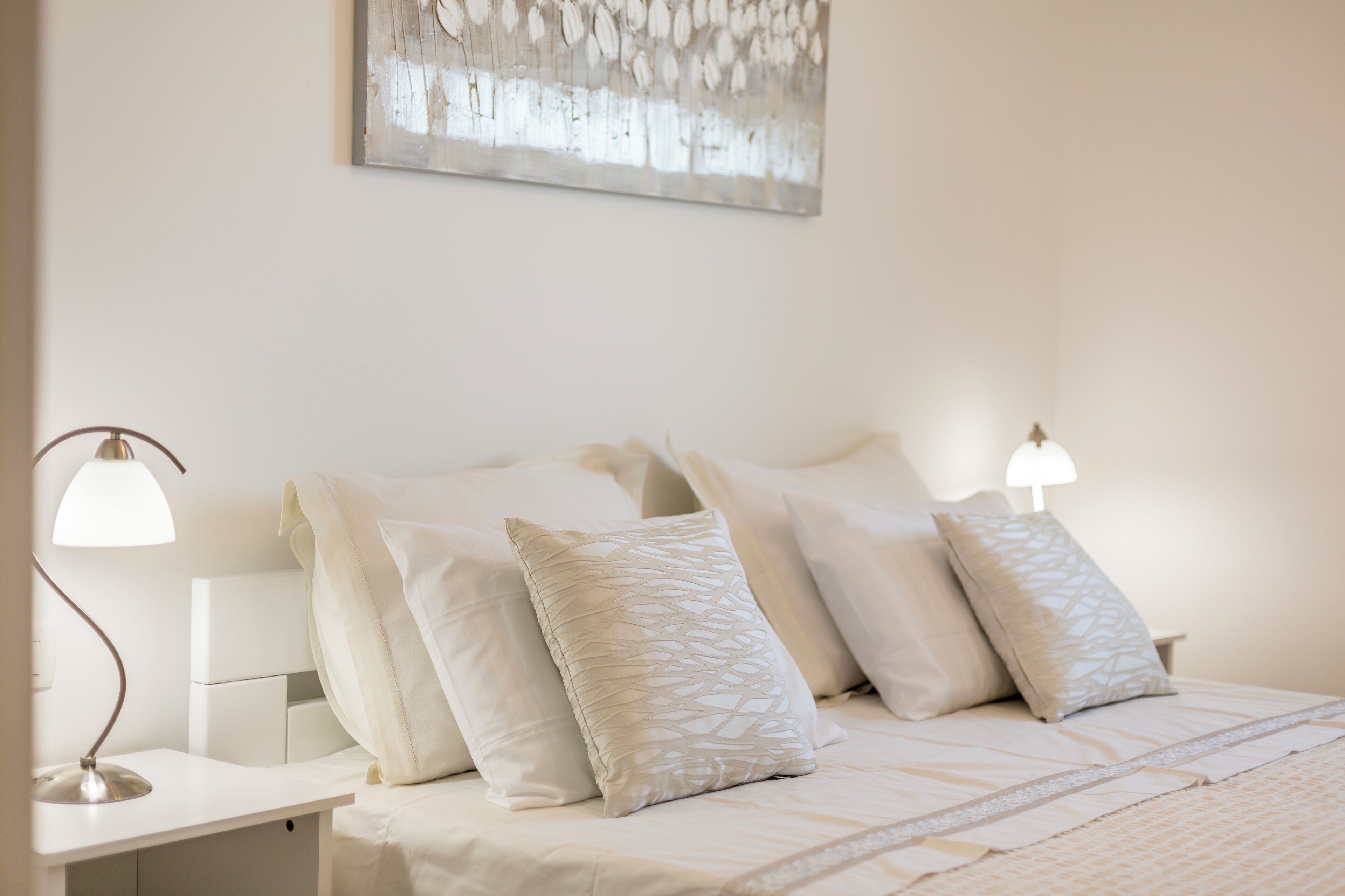 A neatly made bed with white linens and multiple pillows, a bedside table with a lamp, and a wall-mounted abstract art piece.