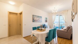 Un área de comedor con una mesa redonda, sillas color turquesa y una mesita de madera con una botella y copas. Hay un sofá y un cuadro en la pared.