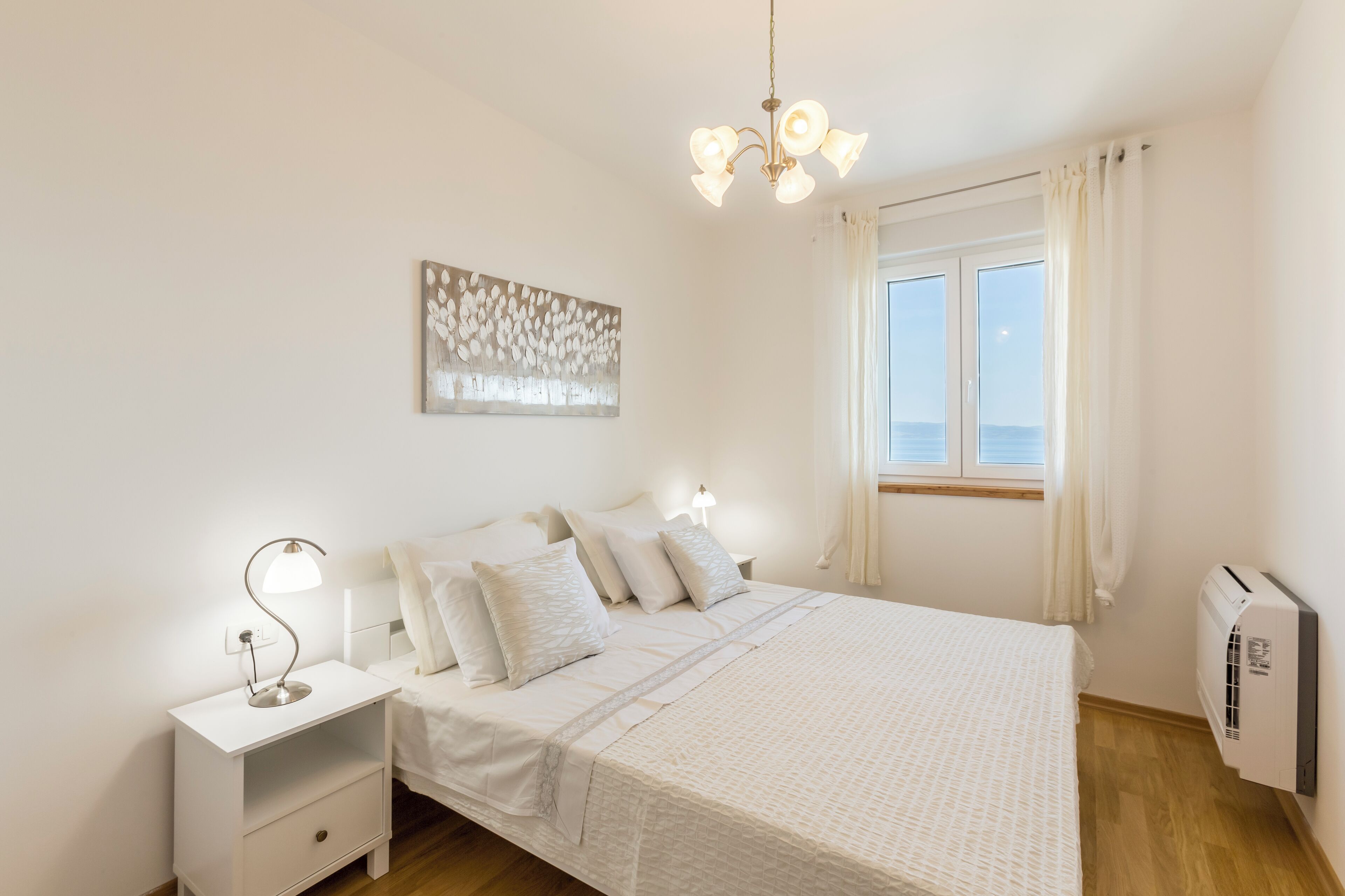 A bedroom with a bed, bedside lamps, a wall art piece, a window with curtains, and an air conditioning unit.