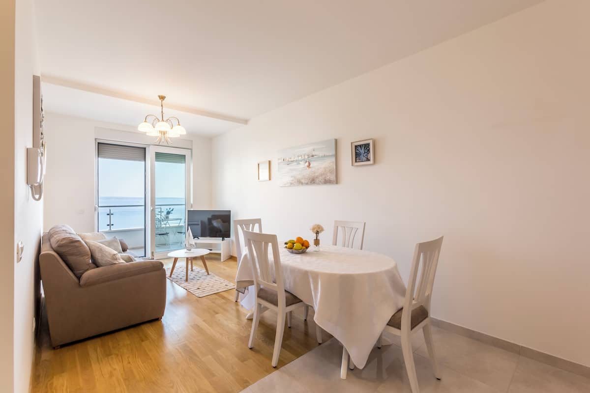 Una sala de estar moderna con sofá, mesa de comedor y sillas. Hay un televisor y una ventana con vista al mar.