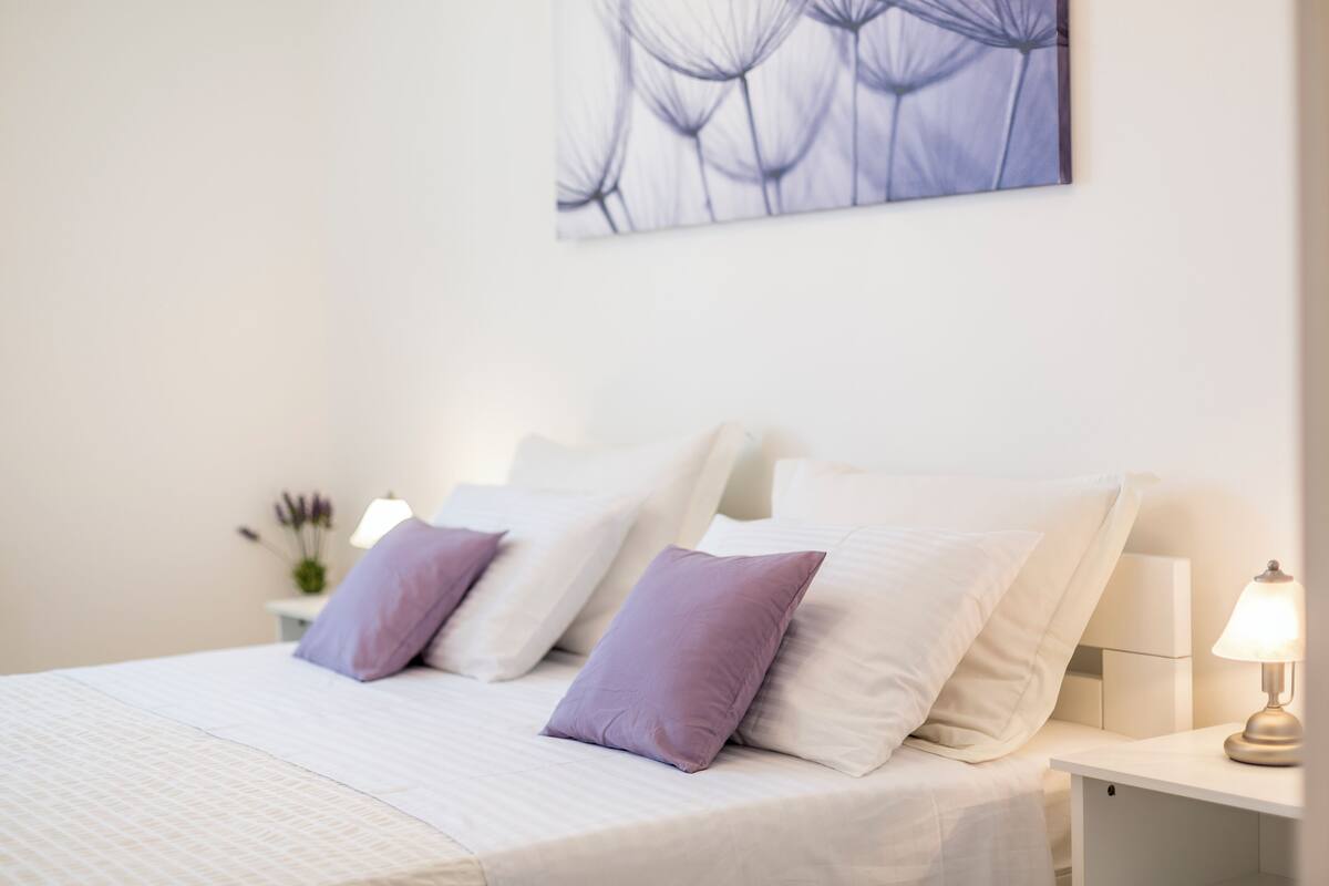 Una cama bien hecha con almohadas blancas y púrpuras, una mesita de noche con lámpara y una obra de arte colgada en la pared.
